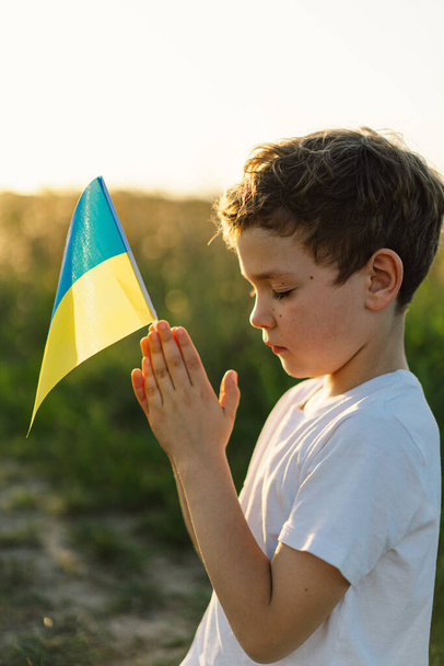 Oekraïense jongen sloot haar ogen en bad om de oorlog in Oekraïne te stoppen in een veld bij zonsondergang. Handen gevouwen in gebed concept voor geloof, spiritualiteit en religie. Oorlog van Rusland tegen Oekraïne. Stop de oorlog - Foto, afbeelding