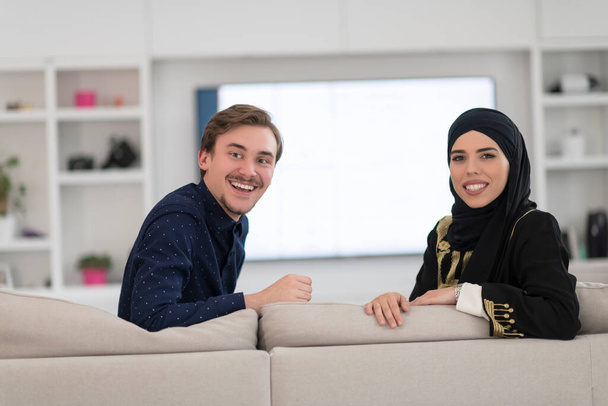 Young Muslim couple woman wearing Islamic hijab clothes sitting on the sofa watching TV together during the month of Ramadan at a modern home.  - Fotografie, Obrázek