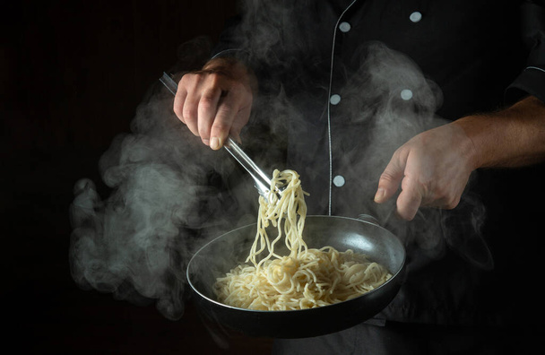 Der Koch kocht Spaghetti in einer heißen Pfanne. Platz für Werbung auf schwarzem Hintergrund. Italienische Nationalküche. - Foto, Bild