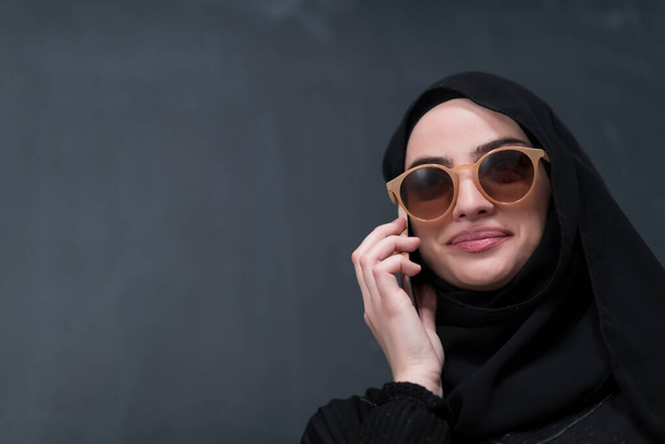 黒の黒板の前にサングラスとヒジャーブの服を着た若い現代のイスラム教徒のビジネス女性.  - 写真・画像