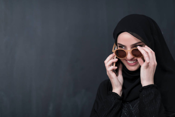 Young modern Muslim businesswoman using smartphones wearing sunglasses and hijab clothes in front of a black chalkboard.  - Foto, Bild