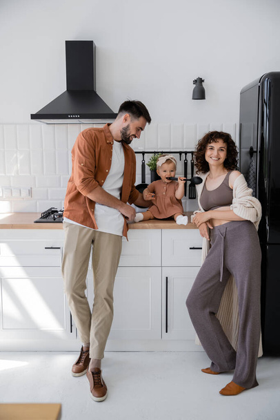 Säugling mit Stirnband hält Schöpfkelle in der Nähe des Mundes, während er auf Küchenarbeitsplatte in der Nähe glücklicher Eltern sitzt  - Foto, Bild