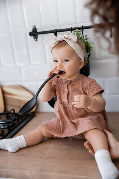 Säugling mit Stirnband hält Schöpfkelle in der Nähe des Mundes, während er auf Küchenarbeitsplatte in der Nähe der Mutter sitzt - Foto, Bild