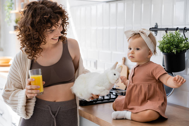 infant girl sitting on kitchen worktop near cheerful mother with glass of orange juice and soft toy   - Foto, immagini