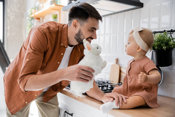 happy bearded man holding soft toy near infant daughter sitting on kitchen worktop  - Photo, Image