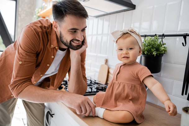happy bearded man touching tiny leg of infant daughter sitting on kitchen worktop  - 写真・画像