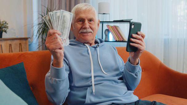家族の予算を計画してる。高齢者の祖父の笑顔お金の現金を数える、自宅で国内の請求書を計算スマートフォンを使用します。楽しい男は収入に満足し、計画休暇のためのお金を節約,贈り物 - 写真・画像