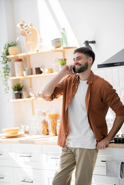 happy bearded man talking on smartphone while standing in modern kitchen  - 写真・画像