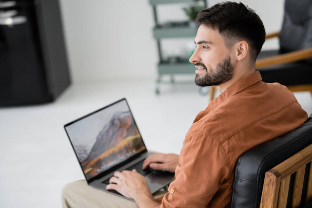 cheerful and bearded freelancer sitting on leather armchair with modern laptop  - Foto, Bild