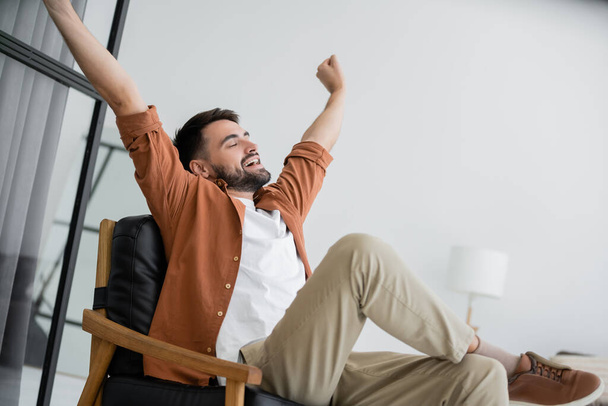 bearded man stretching and yawning while sitting on comfortable leather armchair in living room  - Фото, зображення