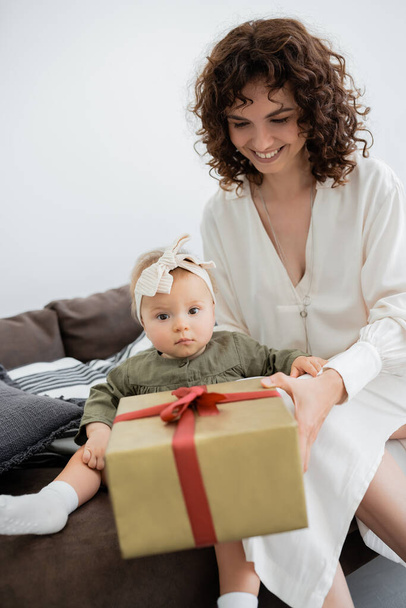 cheerful mother in dress holding wrapped gift box near infant daughter in headband sitting on sofa - Fotografie, Obrázek