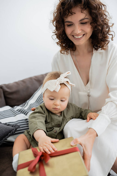örömteli anya kezében csomagolva ajándék doboz közelében csecsemő lánya fejpánt ül a kanapén - Fotó, kép