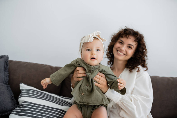 happy mother with curly hair smiling while holding in arms infant daughter in headband   - Fotografie, Obrázek