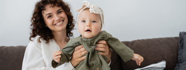 happy mother with curly hair smiling while holding in arms infant daughter in headband, banner - Photo, Image