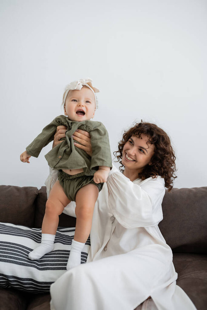 happy mother with curly hair smiling while holding in arms excited infant daughter in  headband  - Foto, Bild