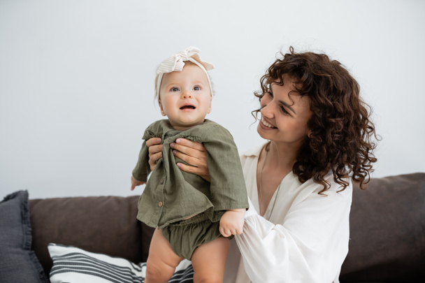 happy mother with curly hair smiling while holding in arms baby daughter in dress and headband  - Fotó, kép