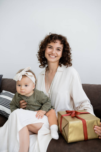 pleased and curly mother in dress holding wrapped gift box near infant daughter in headband - 写真・画像