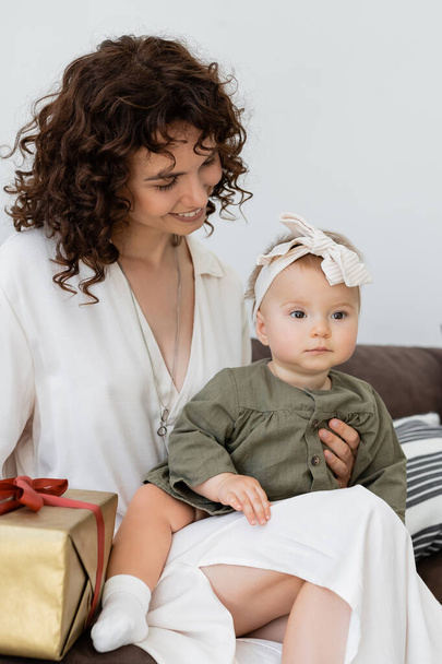happy and curly mother in dress holding wrapped gift box near infant daughter in headband - Photo, Image