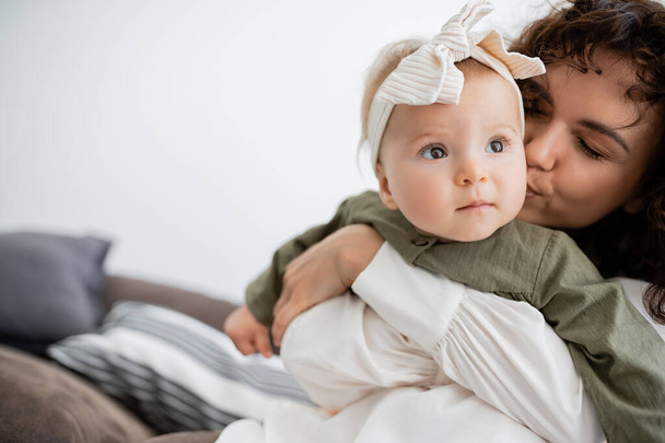 curly mother hugging and kissing cheek of baby girl in headband  - Photo, Image