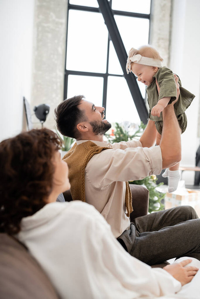 cheerful father holding infant daughter near blurred wife in living room  - Foto, imagen