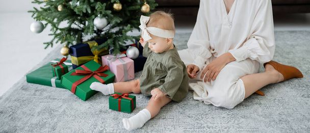 装飾されたクリスマスツリーの近くのギフトボックスを見ている幼児の娘の近くに座っている母親バナー - 写真・画像