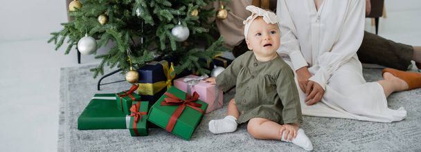 rodina s kojeneckou holčičkou sedí v blízkosti vánočního stromku s dárky, banner - Fotografie, Obrázek