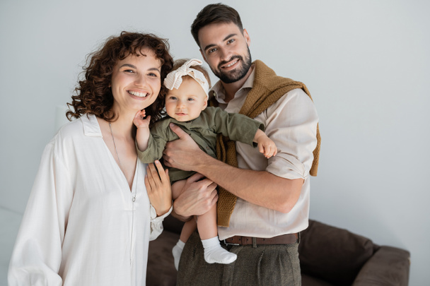 positive bearded man holding in arms baby daughter in headband near curly wife  - Photo, image