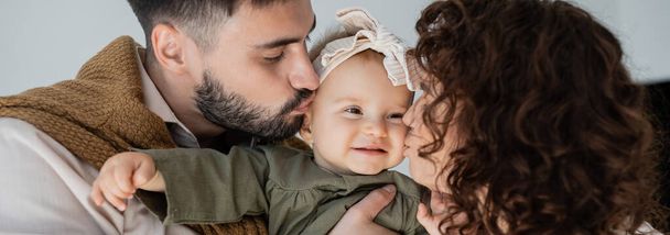 bearded man and curly woman kissing cheeks of happy baby daughter in headband, banner - Фото, зображення