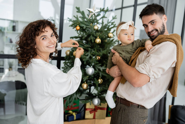 cheerful woman decorating christmas tree near happy husband and infant daughter - Foto, Imagen