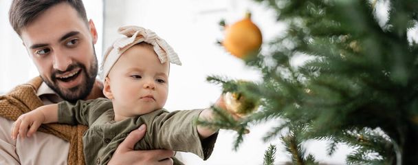 padre emocionado sosteniendo hija bebé cerca del árbol de Navidad decorado, pancarta - Foto, imagen
