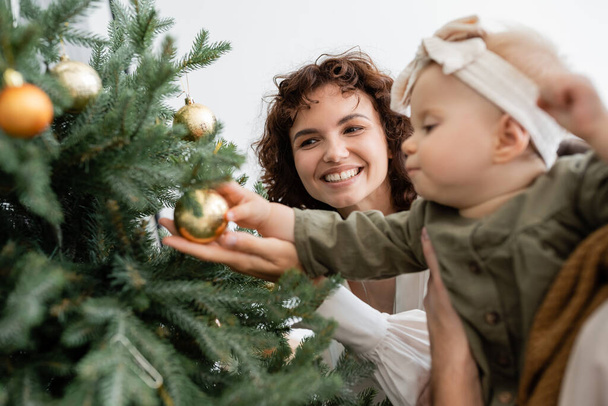 padre che tiene la figlia infante vicino all'albero di Natale decorato e alla moglie allegra - Foto, immagini
