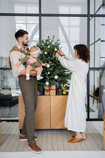 кудрявая женщина украшает рождественскую елку рядом с бородатым мужем и маленькой дочерью  - Фото, изображение