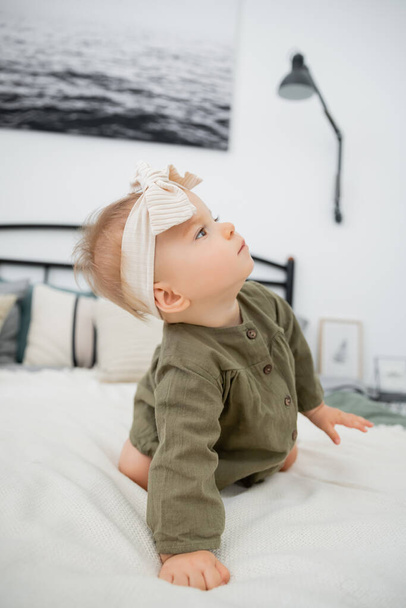 curious baby girl in dress and headband with bow sitting on bed at home - Photo, image