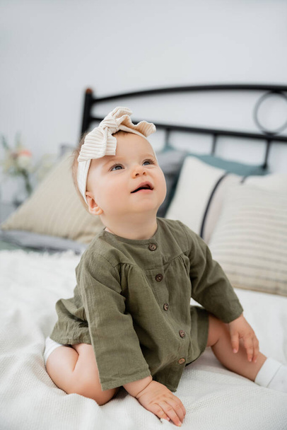 cheerful baby girl in dress and headband with bow sitting on bed at home - 写真・画像