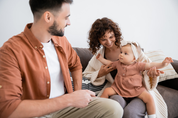 cheerful mother holding infant daughter near bearded husband sitting on couch - Photo, Image