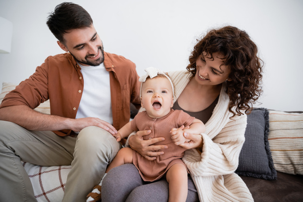 cheerful mother holding amazed infant daughter near bearded husband sitting on couch - Foto, Imagem