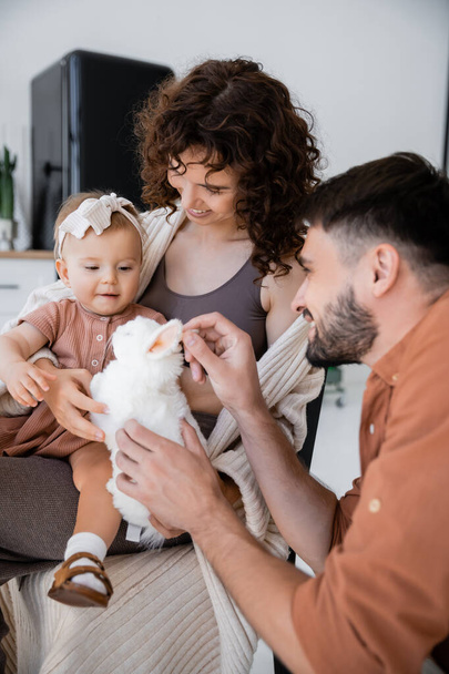 happy bearded man holding soft toy near baby daughter and cheerful wife  - Fotó, kép