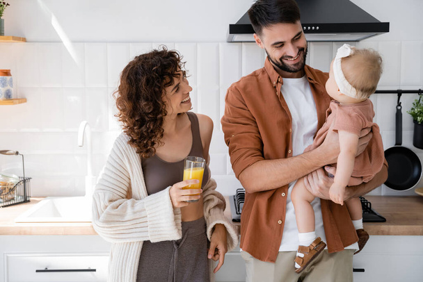 довольный мужчина держит маленькую дочь рядом с кудрявой женой со стаканом свежего апельсинового сока на кухне  - Фото, изображение