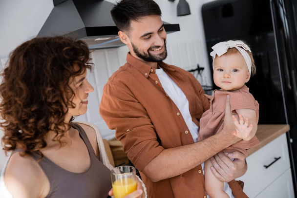 happy man holding baby daughter near curly wife with glass of fresh orange juice in kitchen   - Foto, imagen