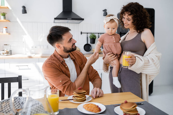 cheerful wife holding in arms infant daughter and bringing orange juice to husband during breakfast  - 写真・画像