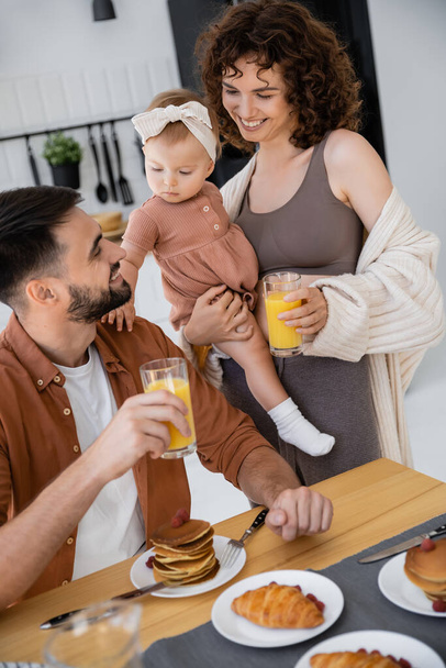glückliche Frau mit Baby im Arm hält Glas Orangensaft neben fröhlichem Ehemann  - Foto, Bild