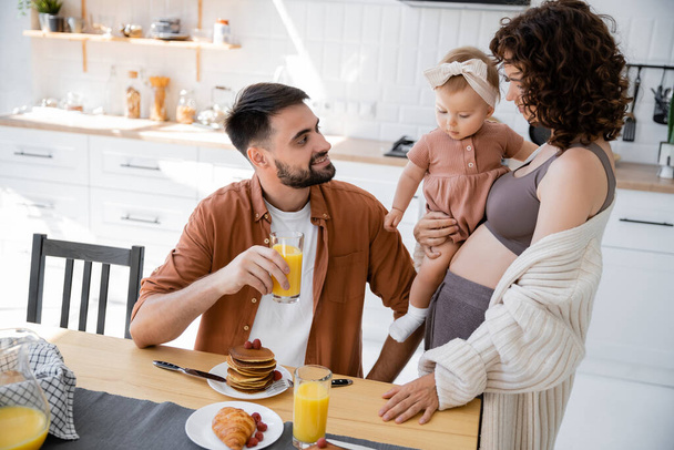 кудрявая женщина с младенцем на руках смотрит на улыбающегося мужа во время завтрака  - Фото, изображение