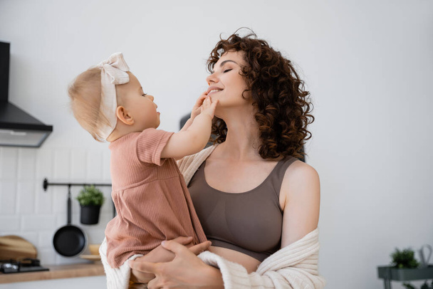 happy woman with curly hair smiling while holding in arms infant baby in headband  - Photo, image