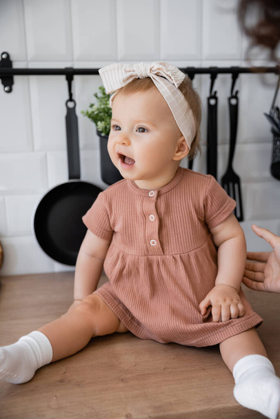 amazed infant girl in headband sitting on kitchen worktop near blurred mother - Foto, afbeelding
