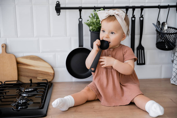 infant girl in headband and pink dress holding ladle near mouth while sitting on kitchen worktop - Foto, Bild