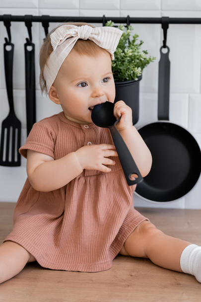 Baby Mädchen mit Stirnband und rosa Kleid hält Schöpfkelle in der Nähe des Mundes, während sie auf der Arbeitsplatte in der Küche sitzt - Foto, Bild