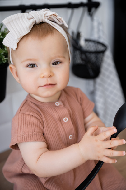 Saç bandı takmış küçük bir kızın portresi ve mutfak atölyesinde otururken elinde kepçe tutan pembe bir elbise. - Fotoğraf, Görsel