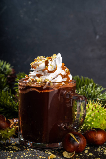 Glass cup with dark chestnut hot chocolate or choco latte, with whipped cream, caramel sauce and roasted chestnuts, over dark table copy space - Zdjęcie, obraz