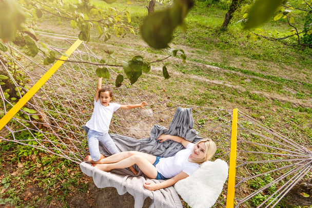 Glückliche Mutter und Tochter relaxen zusammen in einer Hängematte im Garten an einem sonnigen Sommertag. Familie spielt in Hängematte. - Foto, Bild