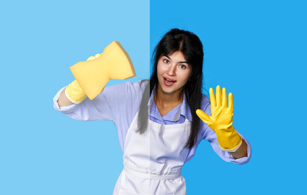 young confident woman in rubber gloves and cleaner apron cleaning glass with yellow sponge on blue background. - Photo, Image